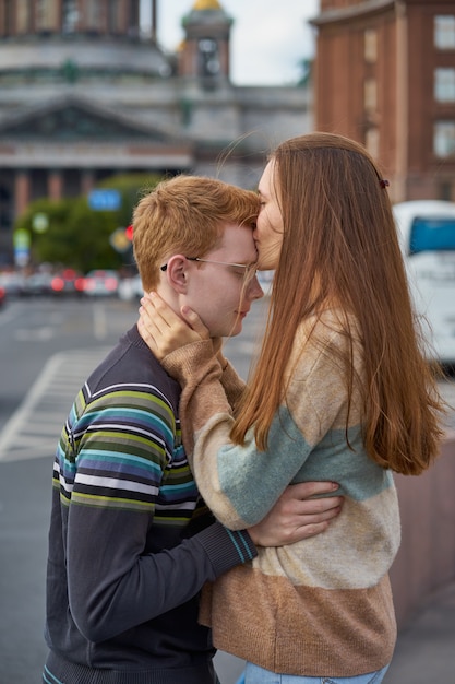 Red-haired woman kisses a man on the top of her head, a woman with long dark thick hair in sweater