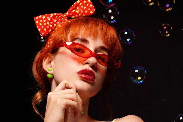 Red-haired woman with a bright headband and red glasses on a black background.