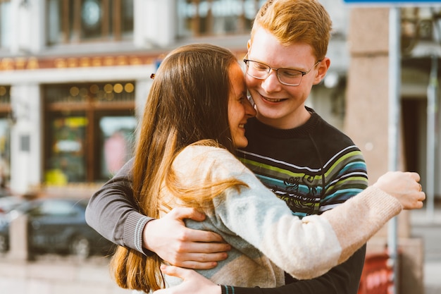 A red-haired young man embraces young woman with long dark hair, they laugh 