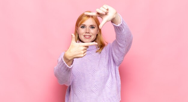 Red head pretty woman feeling happy, friendly and positive, smiling and making a portrait or photo frame with hands