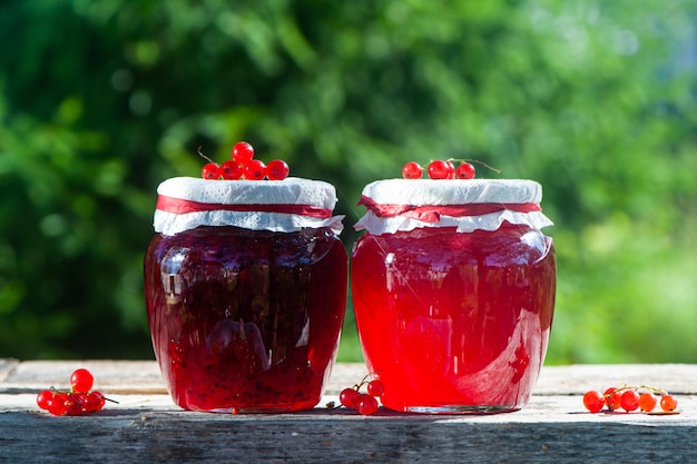 Red juicy berries of red currants and jars of berry jam on a wooden table on a green background of the garden harvest and cooking theme