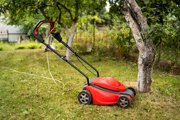 Red lawn mower outdoor in the backyard green grass and fruit trees background Gardening concept