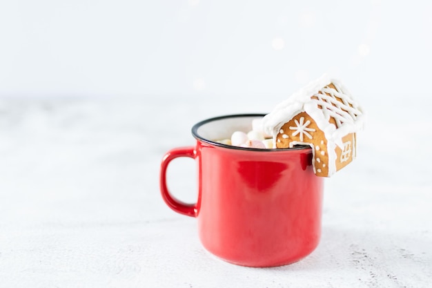 Red mug of hot chocolate with marshmallow and homemade gingerbread small house for decoration