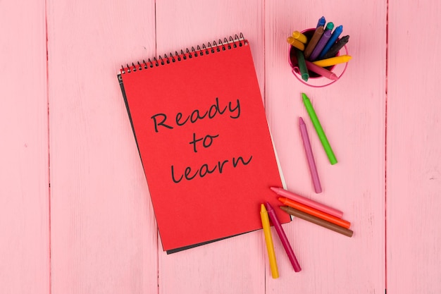 Red note pad with text Ready to learn and crayons on pink wooden table