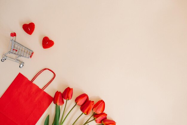 Red paper shopping bag a bouquet of tulips and a shopping cart Holiday discounts for Valentines Day with space copying A frame of objects on a beige background