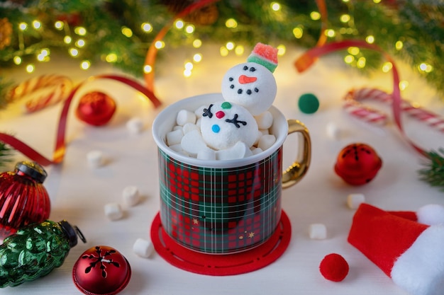 A red plaid mug with Snowman and marshmallows on a background of Christmas decorations and fir branches