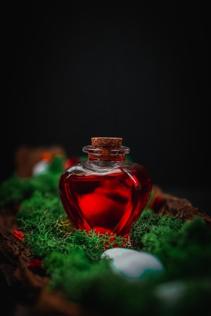 Red poison in a heartshaped bottle lies in the bark of a tree with moss and stones on a black