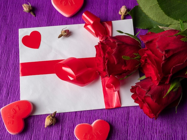 Red roses red hearts next to rosebuds and card with ribbon on purple background