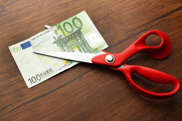 Red scissors cut money on wooden background Financial concept