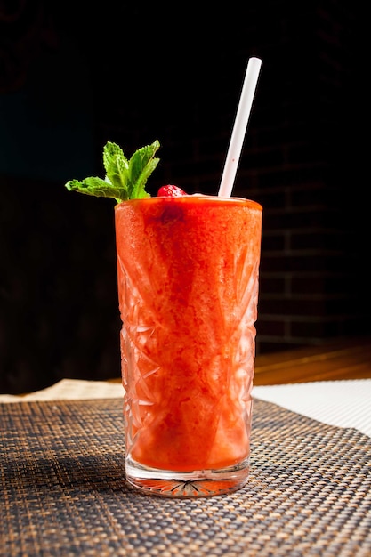 Red strawberry alcoholic cocktail with mint and straw. Alcoholic or non-alcoholic cocktail in a long, tall and narrow clear glass with a dark red liquid and syrup and mint on a dark background