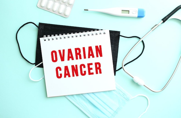 The red text OVARIAN CANCER is written on a white sheet of notepad that lies on a blue background next to a thermometer and medical face masks