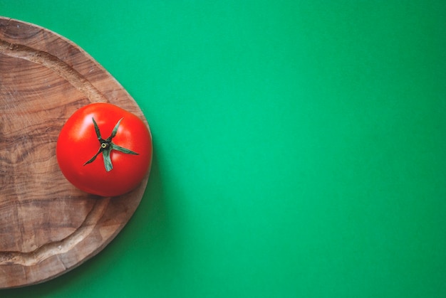 Red tomato on a wooden cutting board on a green surface. Taken by a camera from above.