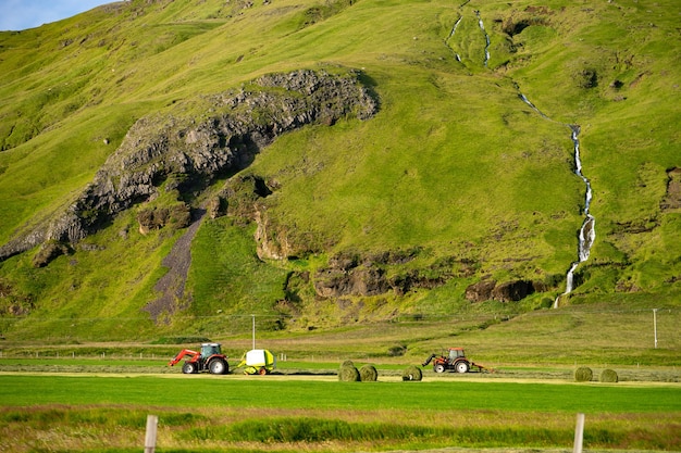 Red Tractor collecting stack of grass on green field with beautiful river going by the mountain on background.