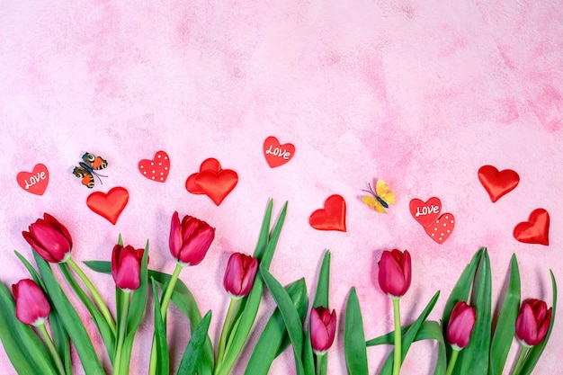 Red tulips, red hearts and fluttering butterflies on a pink textured background with a copy of space