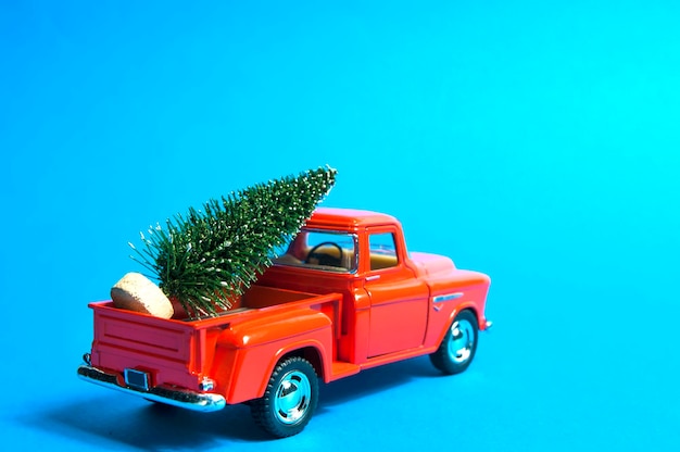 A red vintage pickup truck carries a green Christmas tree on a blue background, rear side view, selective focus.