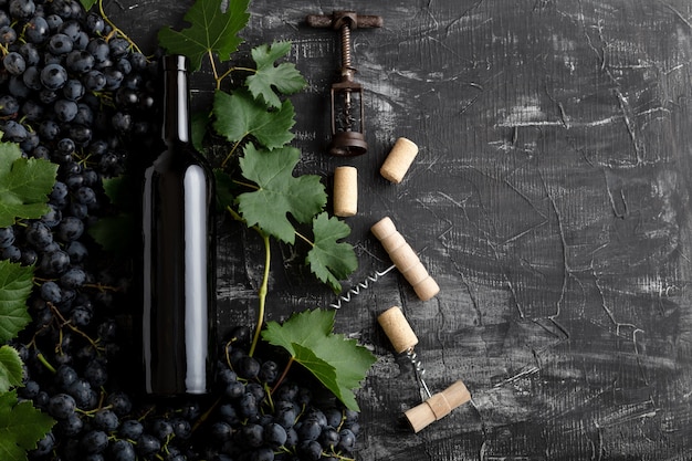 Red Wine bottle grape bunches with leaves and vines corkscrew on dark rustic concrete background. Flat lay wine composition Red Wine bottle on black stone table with copy space.