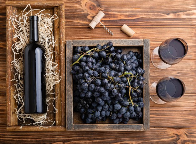 Red Wine composition on brown wooden table. Top view. Red wine bottle corkscrew corks wine glasses black ripe grapes in box on wooden table.