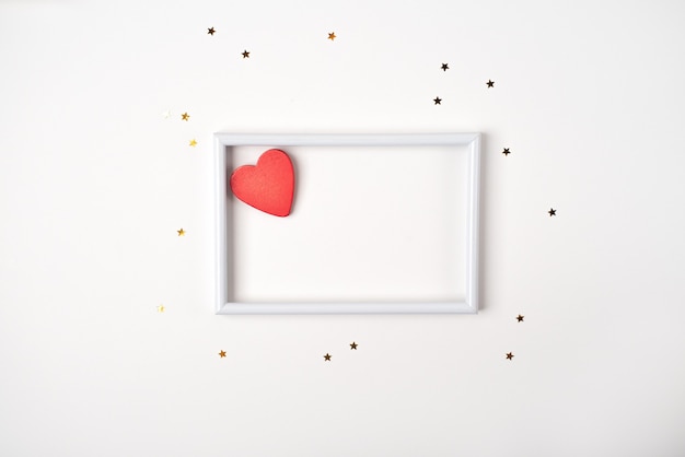 Red wooden heart in the white frame and little golden stars on the light (white) background