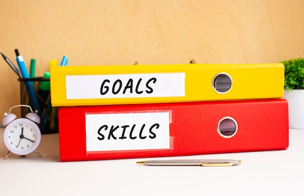 Red and yellow folders lie on the office table next to the clock and pen Inscriptions on GOALS and SKILLS folders