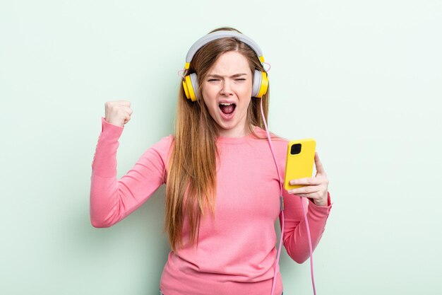 Photo redhair woman shouting aggressively with an angry expression headphones and smartphone concept