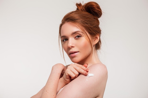 Redhaired beautiful young woman with clean facial skin applies cream on her shoulder in the studio on a white background Beauty and body skin care