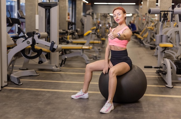 Redhaired cheerful fit woman sits on a fitball in a modern gym Shows thumb up