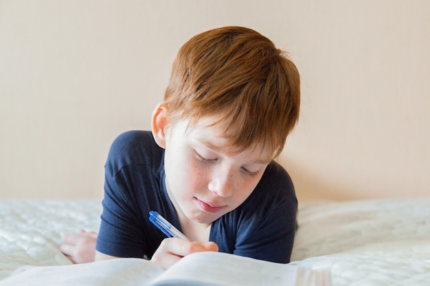 A redhaired European boy at home on the bed writes homework in front of him an open notebook