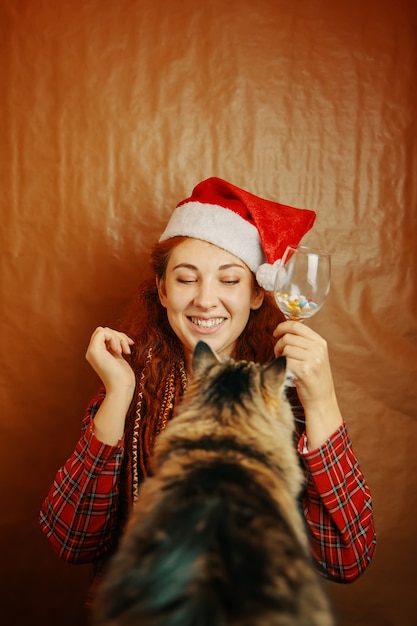 Redhaired female in santa claus hat hold glass of pills and play with fluffy cat smiling girl with d...