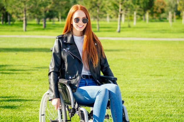 Redhaired ginger woman feeing happy,she sitting wheelchair,wearing sunglasses and warm leather jacket in summer park.