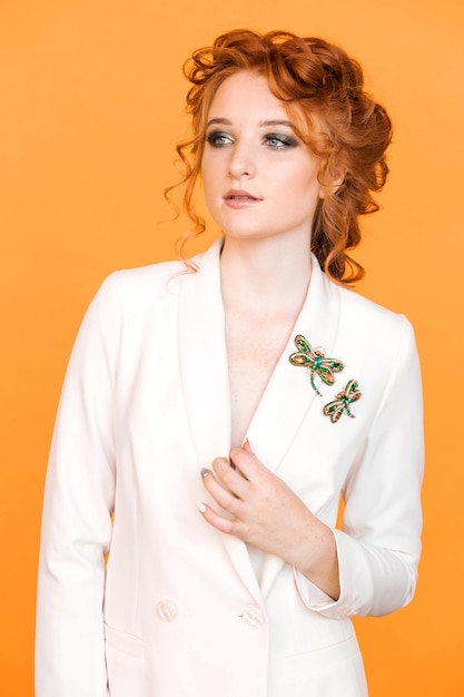 Redhaired girl posing on an orange background in a white jacket with a brooch in shape of dragonfly with green gemstone imitation