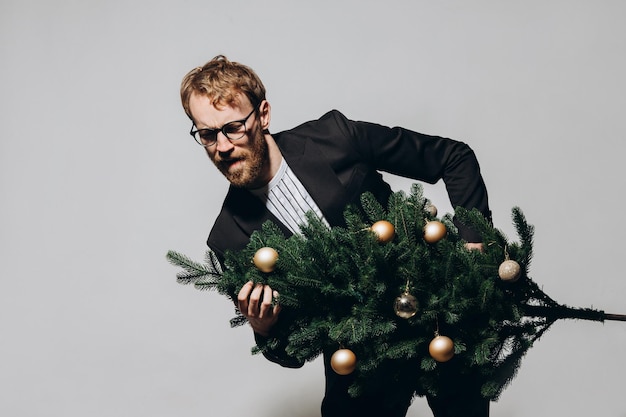 Redhaired handsome man in a black jacket and glasses dances with a Christmas tree in his hands New Year party concept