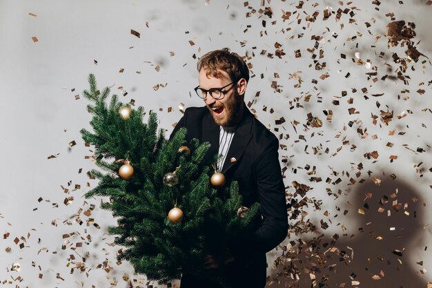 Redhaired handsome man in a black jacket and glasses dances with a Christmas tree in his hands New Year party concept