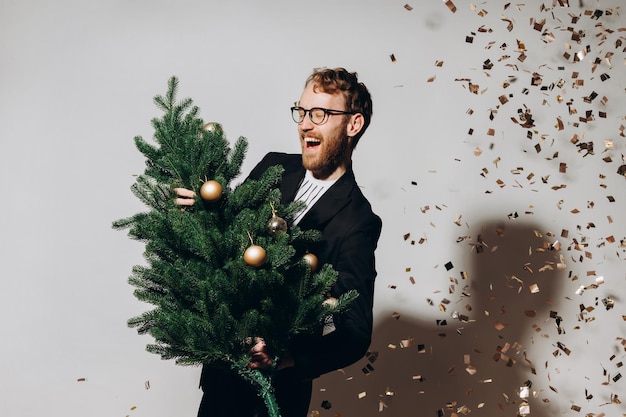 Redhaired handsome man in a black jacket and glasses dances with a Christmas tree in his hands New Year party concept