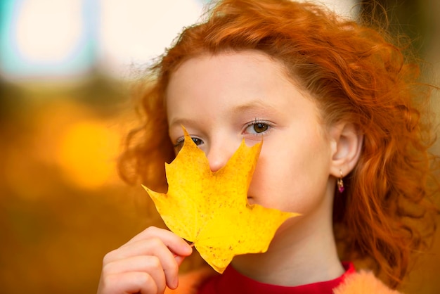 Redhaired little girl closes one eye with a yellow maple leaf Sad child on an autumn day closeup