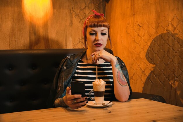 Redhaired pin up styled woman checking her smartphone while having a creamy coffee in a bar