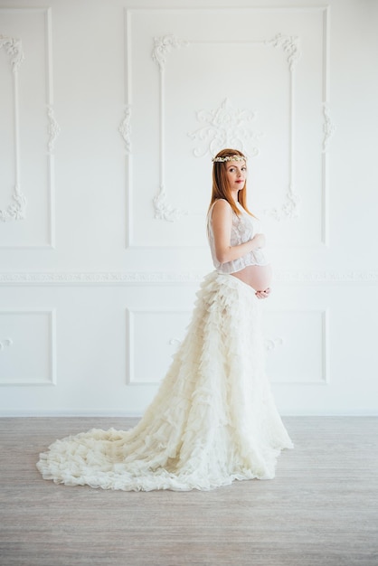 Redhaired pregnant young girl in a white dress near the white wall