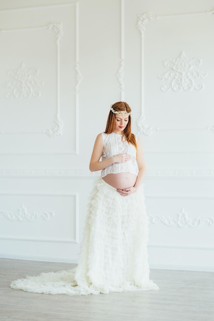 Redhaired pregnant young girl in a white dress near the white wall