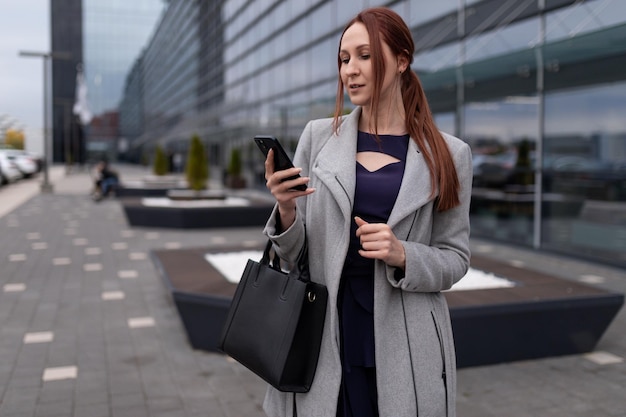 Redhaired woman in business clothes looks at the phone near the office building