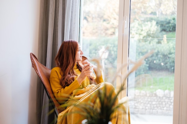 Redhaired woman sitting on the chair by the window with a cup of coffee looking thoughtful Concept Tranquility rest relaxation