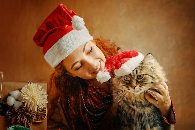 Redhaired woman with dreadlocks in christmas hat holds fluffy tabby cat in santa claus hat next to g...