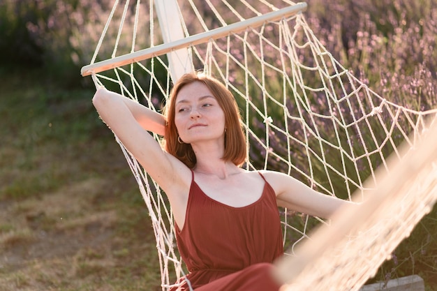 A redhaired young girl without makeup is resting on a hammock in a lavender field Summer vacation and travel time