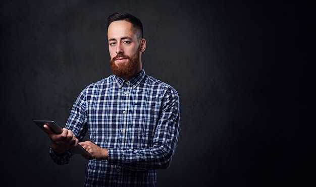 Redhead bearded male dressed in a blue fleece shirt holds tablet PC on grey background.