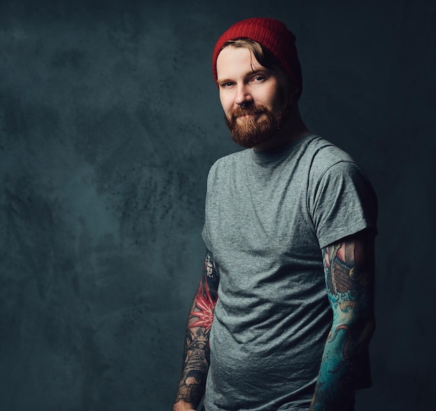 Redhead bearded male with tattoos on his arms posing over grey background.