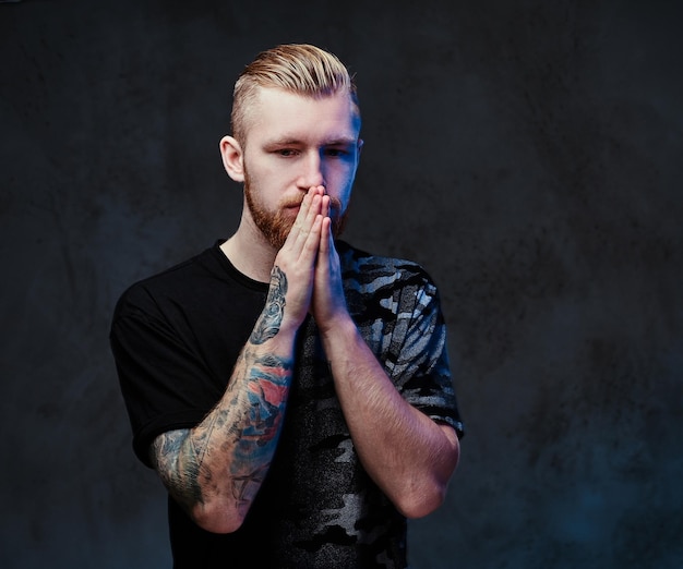 Redhead bearded young male with tattoos on arms, dressed in a black t shirt over dark grey background.