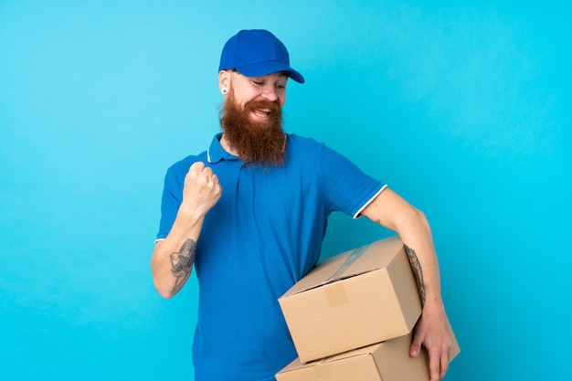 Redhead delivery man over isolated blue celebrating a victory