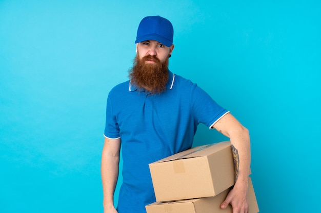 Redhead delivery man over isolated blue wall with sad expression