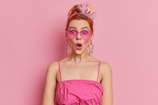  redhead fashionable woman stares surprisingly  keeps mouth opened reacts on amazing news wears trendy sunglasses and dress isolated over pink wall. Style concept