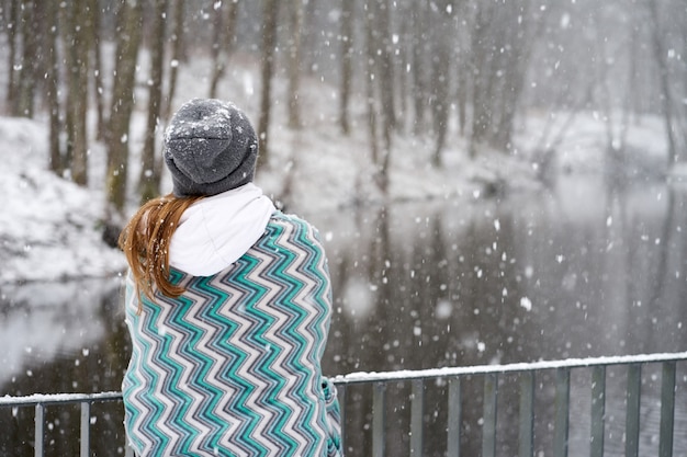 Redhead girl in grey hat, covered in blue patterns plaid enjoying snowfall standing backfront on bridge near river and looking at river