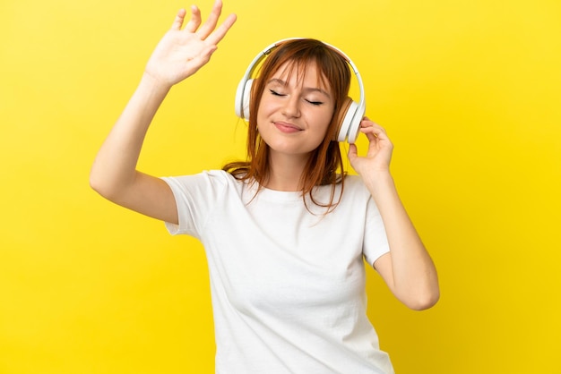 Redhead girl isolated on yellow background listening music and dancing