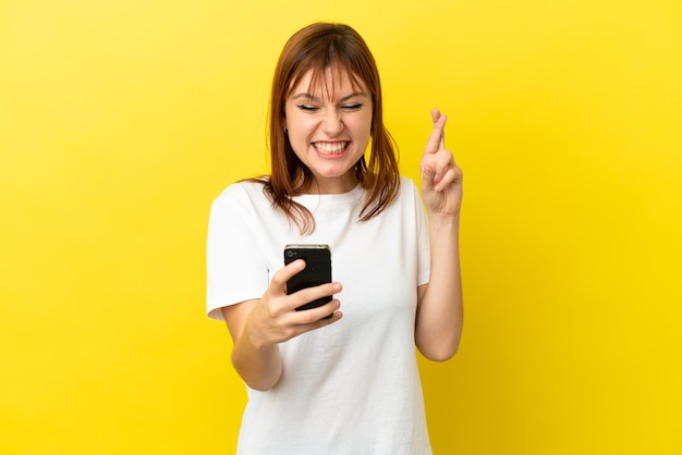 Redhead girl isolated on yellow background using mobile phone with fingers crossing